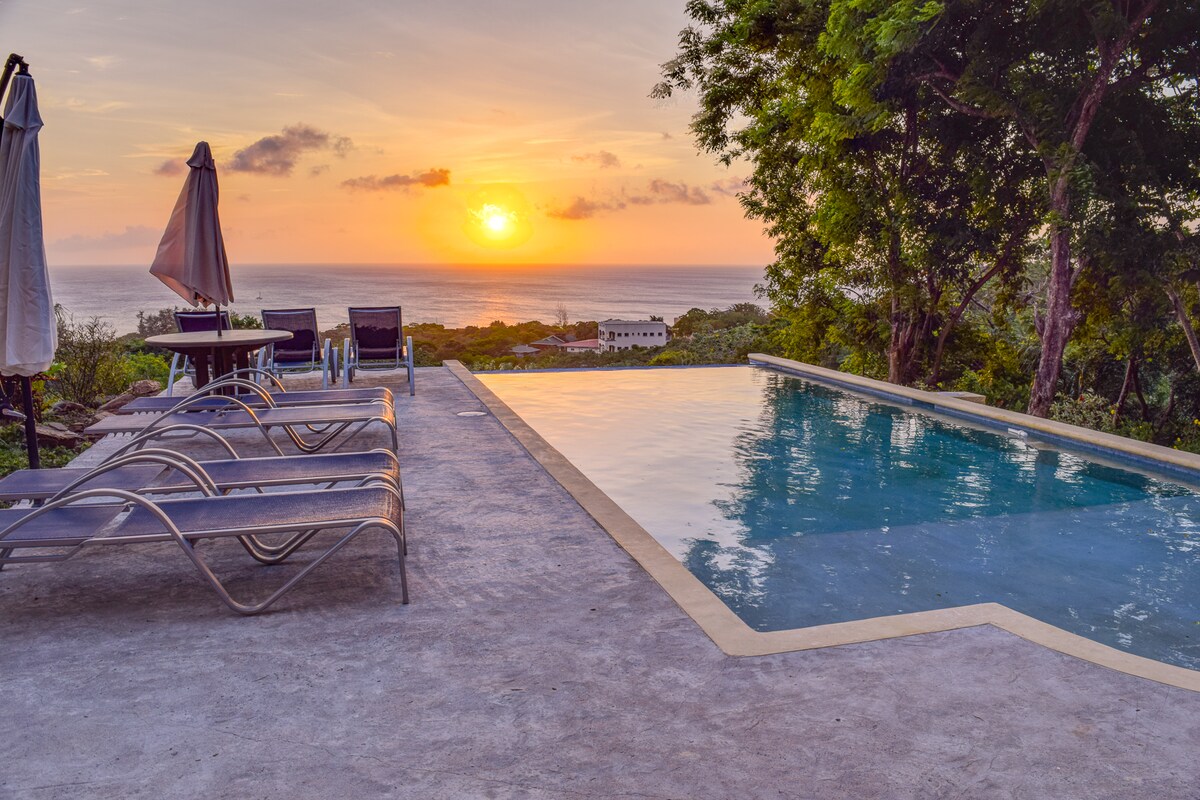 A serene outdoor area showcases a saltwater pool reflecting the vibrant colors of a sunset over the Caribbean Sea. Lounge chairs with umbrellas are positioned nearby, offering a scenic view of the horizon and lush surroundings.