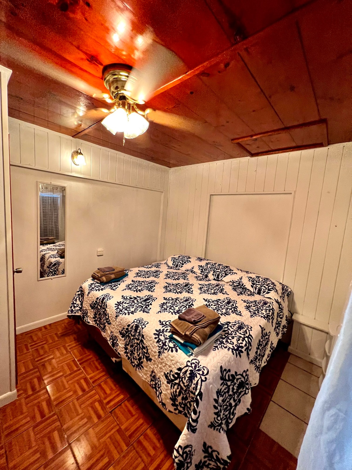 A spacious sleeping area features a large bed covered with a blue and white patterned blanket. Two folded towels are neatly placed at the foot of the bed. A mirror on the wall reflects soft lighting from the ceiling fan, while wooden paneling adds warmth to the room.