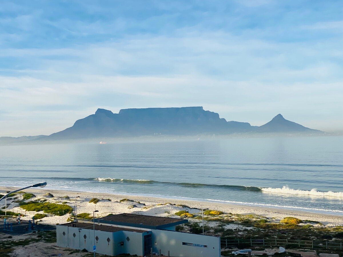 A serene view of Table Mountain and its surrounding peaks is captured against a tranquil ocean backdrop. Soft waves gently lap at the coastline, while the early morning light casts a hazy glow over the scene, highlighting the natural beauty and calmness of the area.