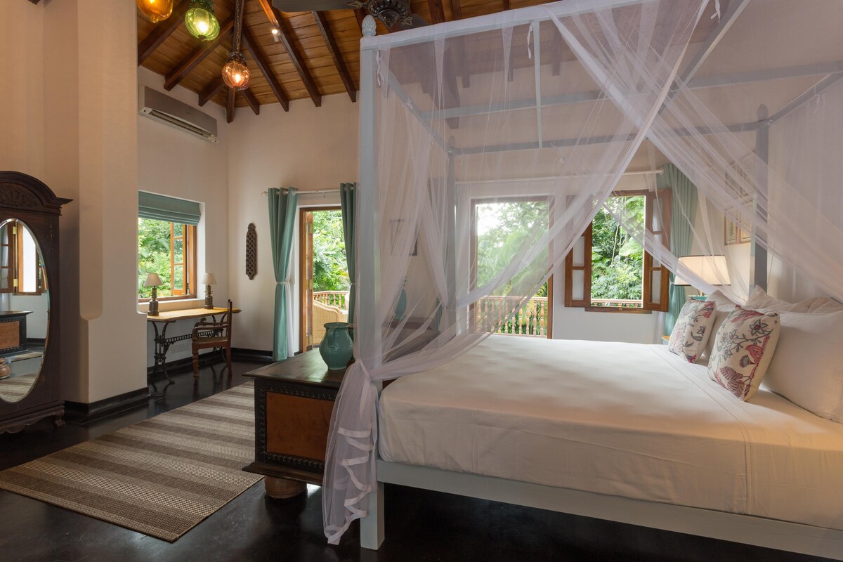 A spacious bedroom features a canopied bed adorned with decorative pillows. Natural light filters through opening windows, illuminating a cozy seating area with a desk and chair. An antique mirror and a stylish rug enhance the room's inviting atmosphere, alongside lush greenery visible outside.