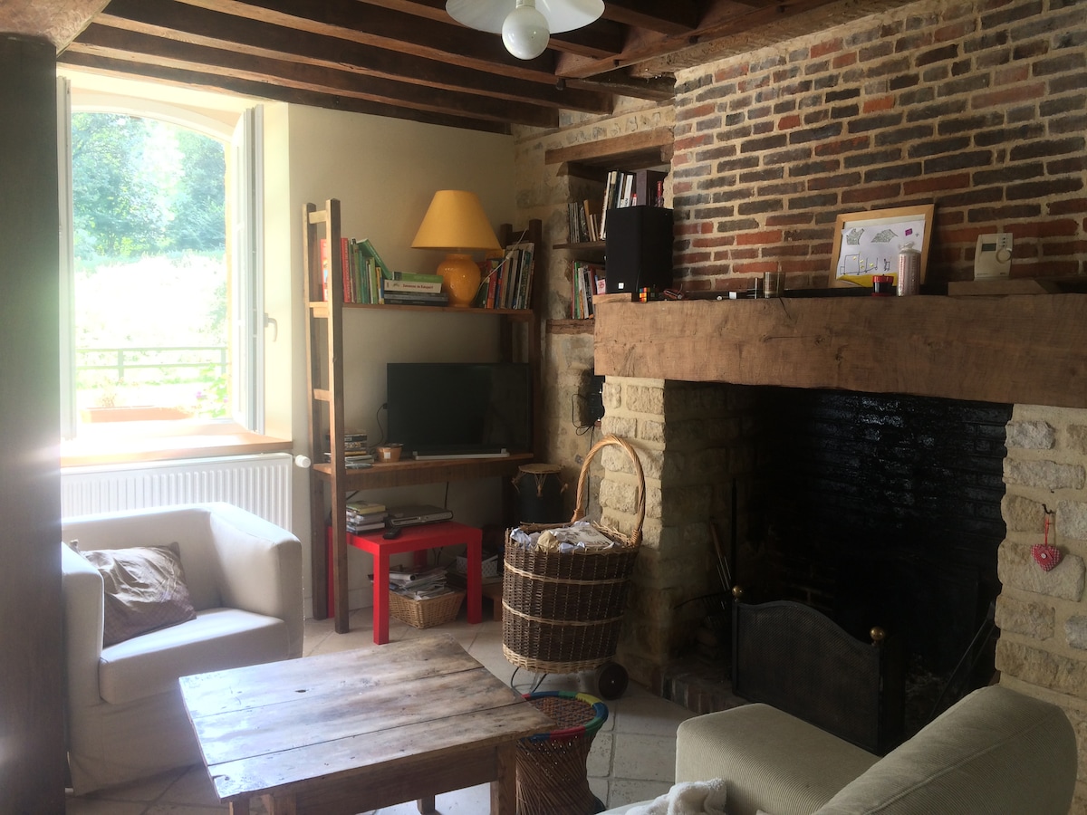 The living area features exposed wooden beams and a rustic stone fireplace. A cozy seating arrangement includes two armchairs and a wooden coffee table. A bookshelf is present, along with a small television. Natural light fills the space through a large window with a view of greenery.