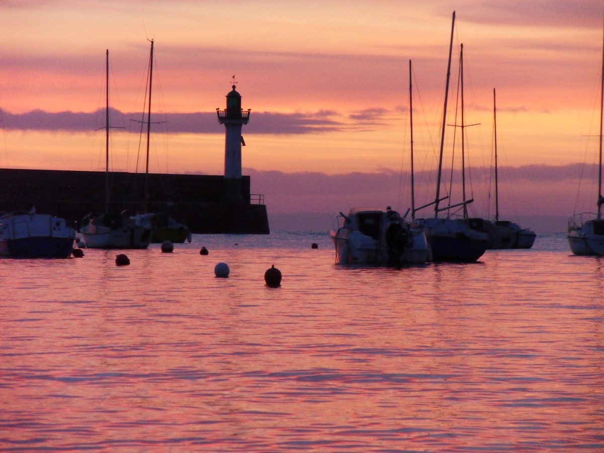 A calm harbor scene is depicted at sunset, featuring various boats gently bobbing on the water. A lighthouse stands tall in the background, silhouetted against a vibrant sky painted in shades of pink and orange. The water reflects the colors, creating a serene atmosphere.