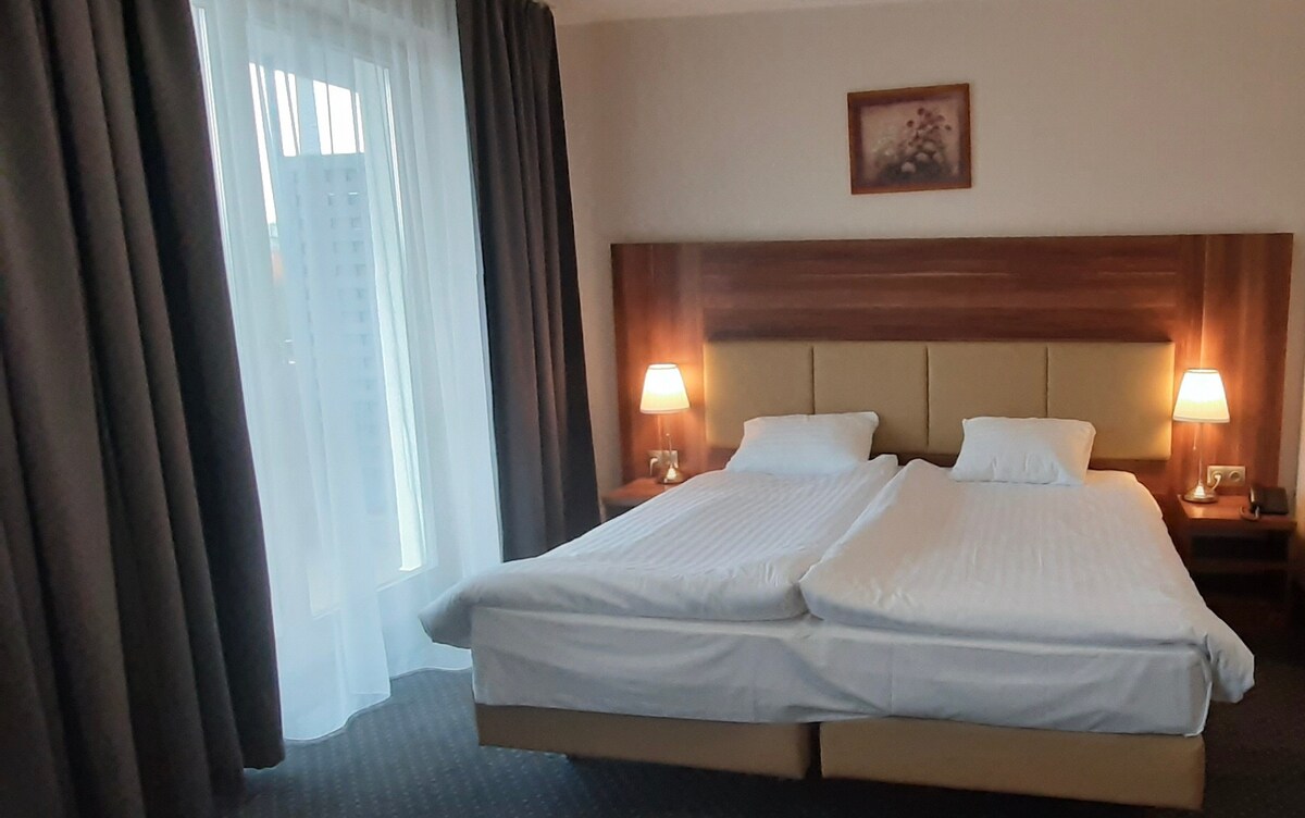 A spacious sleeping area features a large bed configured with two separate mattresses. Soft curtains frame a balcony door, allowing natural light to fill the room. Bedside tables with lamps provide ambient lighting, and a decorative wall art piece adds a subtle touch.