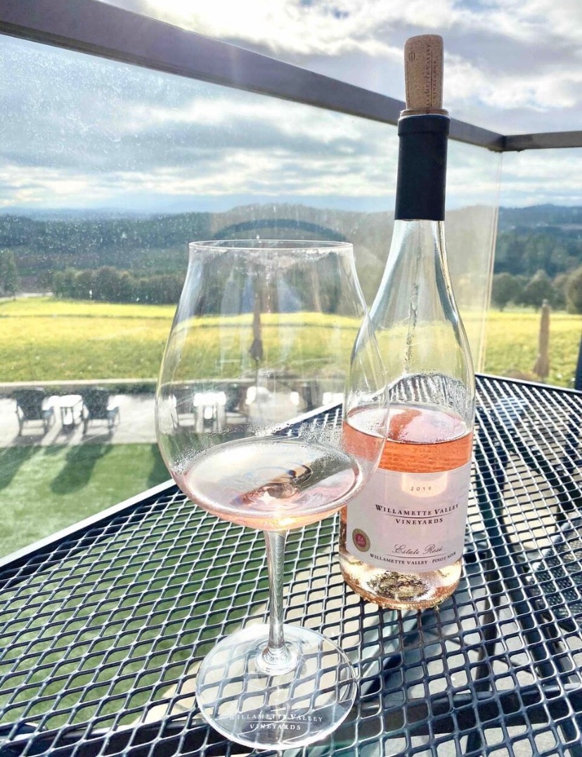 A glass of rosé wine sits on a textured metal table, accompanied by a bottle labeled 'Willamette Valley Vineyards'. A scenic view of rolling green hills and distant mountains is visible in the background, with soft clouds scattered in the sky.