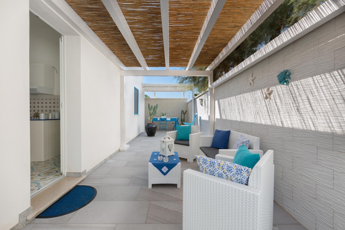 A spacious outdoor area is featured, equipped with a lounge set featuring white wicker chairs and cushions in shades of blue. A small table is positioned centrally, and the space is partially shaded by bamboo overhead, creating a relaxed atmosphere. A kitchenette is visible in the background.