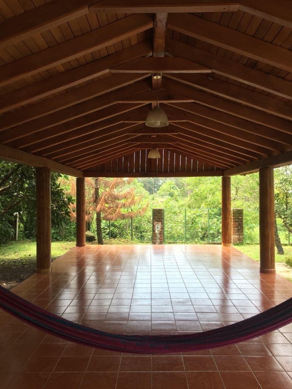 A spacious covered terrace features high wooden ceilings and tiled flooring. Two hanging light fixtures provide illumination. A colorful hammock is suspended, inviting relaxation. The terrace opens to lush greenery, creating an expansive view of the surrounding nature.