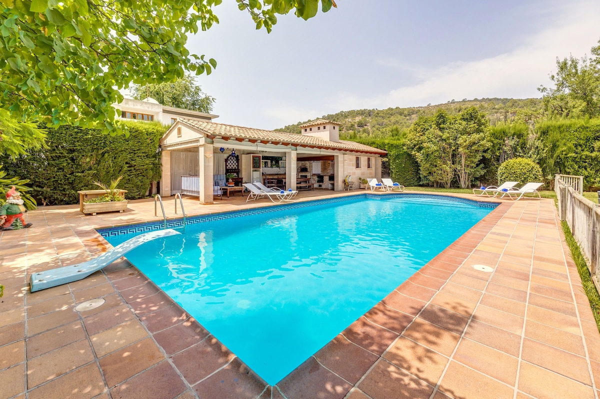 An outdoor area is presented with a large swimming pool surrounded by a spacious sun terrace. Sun loungers are positioned nearby, and a covered outdoor kitchen features a barbecue. Lush greenery and trees flank the area, contributing to a serene and private environment.