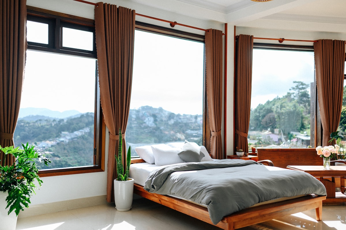 A spacious bedroom features a large wooden bed with a neutral grey comforter. Floor-to-ceiling windows provide panoramic views of the surrounding mountains, while plants in white pots add a touch of greenery to the bright and airy space.