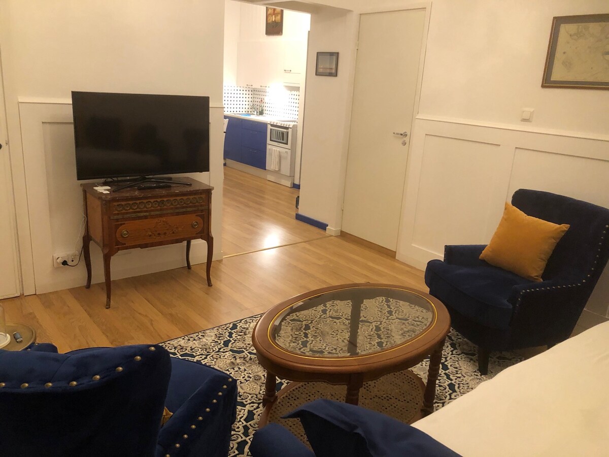The living area features a wooden coffee table surrounded by two blue upholstered chairs with yellow cushions. A television is positioned on a vintage-style cabinet. Light hardwood flooring complements the neutral-colored walls, leading to a view of the kitchen area in the background.