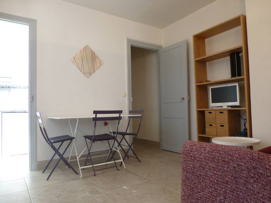 Étagère, télé, ventilo, table et 4 chaises disponibles, cuisine à gauche, couloir menant à la chambre et à la salle de bain à droite.