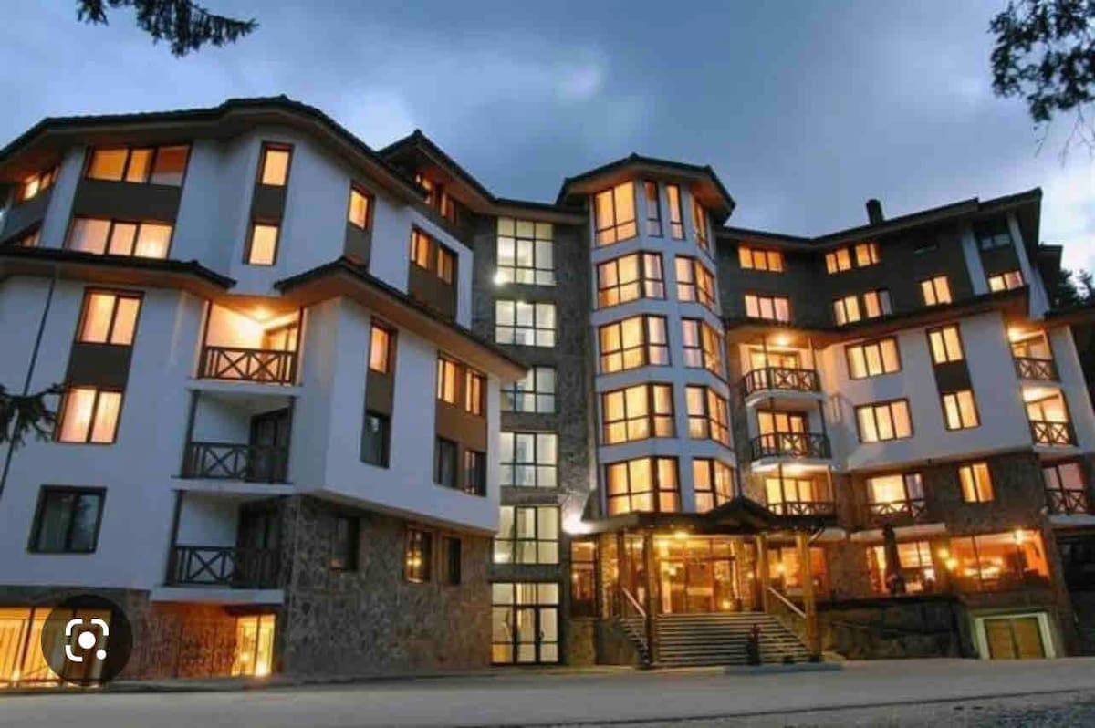 A multi-story building is showcased, featuring a mix of stone and modern architecture. Warm light emanates from numerous windows, creating a welcoming ambiance. Balconies with wooden railings are visible on upper levels. The entrance is framed by a wide staircase leading up to the main door.