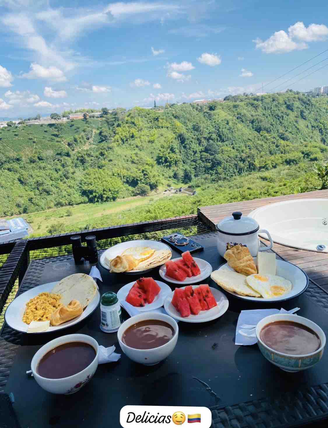 A dining table is set outdoors, featuring various plates with food items, including fruit slices and pancakes. Two bowls of coffee are positioned on the table, with a scenic green hillside visible in the background under a bright blue sky.