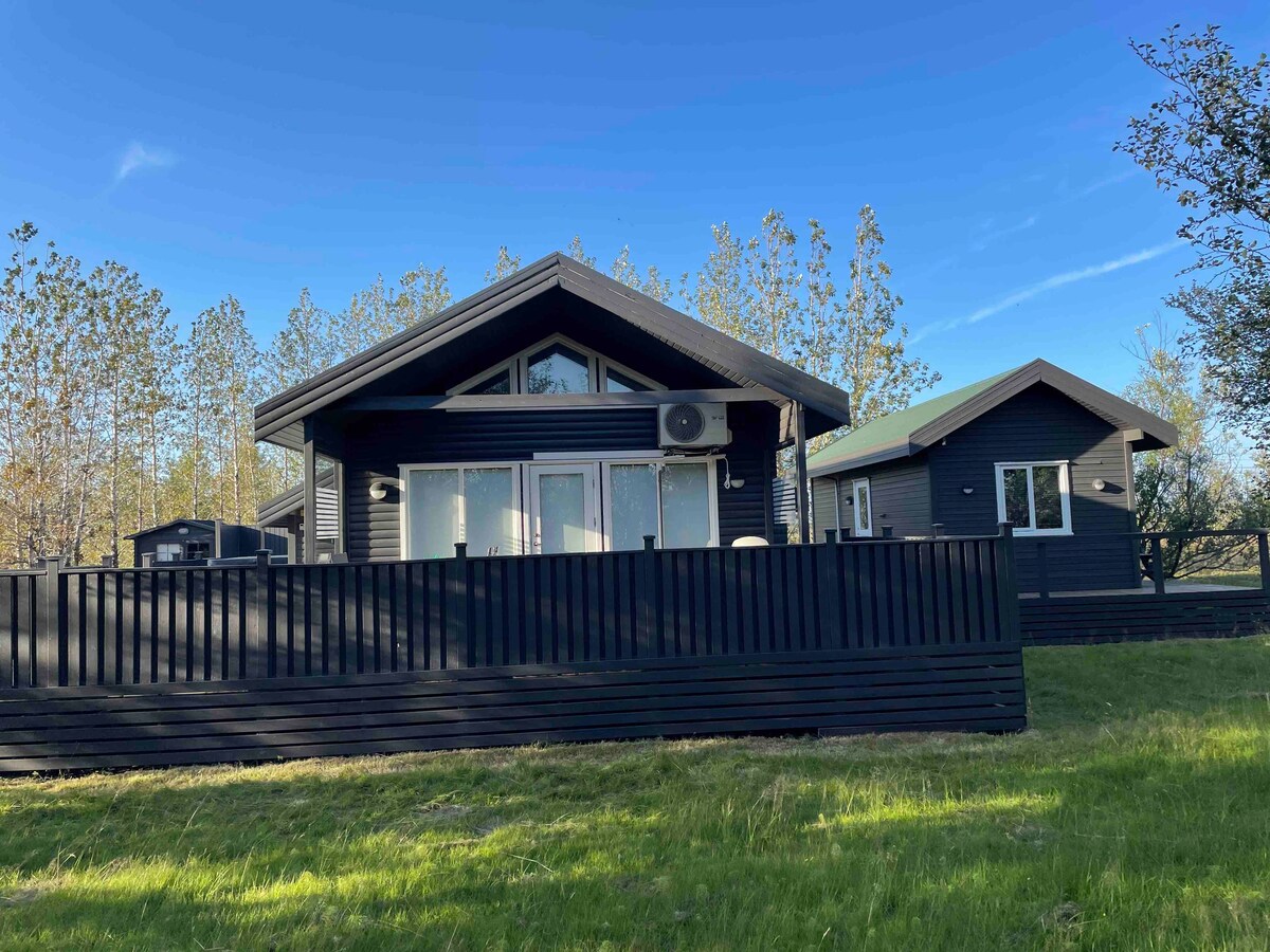 The exterior of a black wooden cottage is framed by lush green grass. Large windows allow ample natural light, while a neighboring smaller house is visible to the right, both surrounded by trees. The clear blue sky adds to the serene environment.