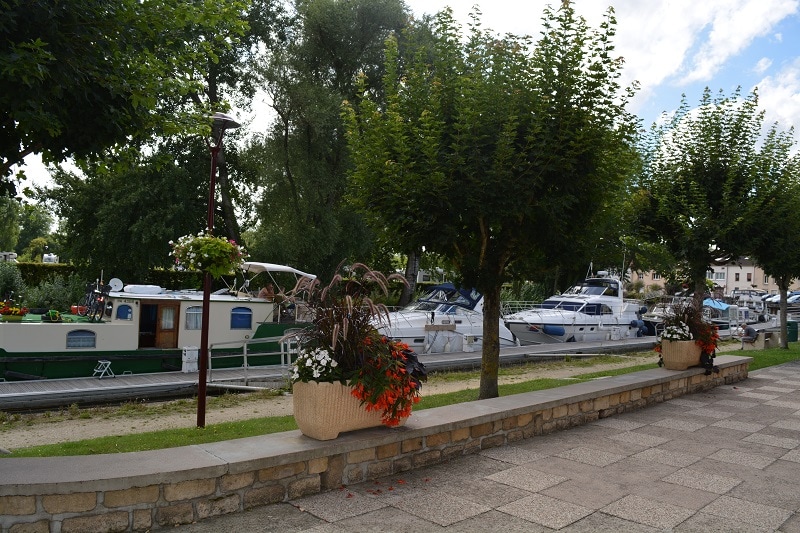 A serene riverside view features moored boats alongside a tree-lined pathway. Decorative planters filled with colorful flowers add splashes of color to the scene. Lush greenery surrounds the area, creating a peaceful atmosphere ideal for relaxation.