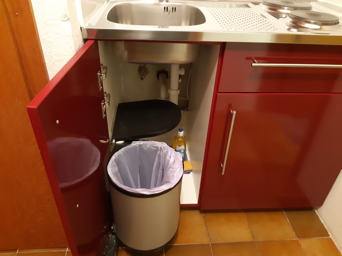 A compact kitchen space features a modern cabinet with a red finish. The open cabinet reveals a trash bin with a liner and cleaning supplies on a shelf above. A stainless steel sink and stove top are visible on the countertop.