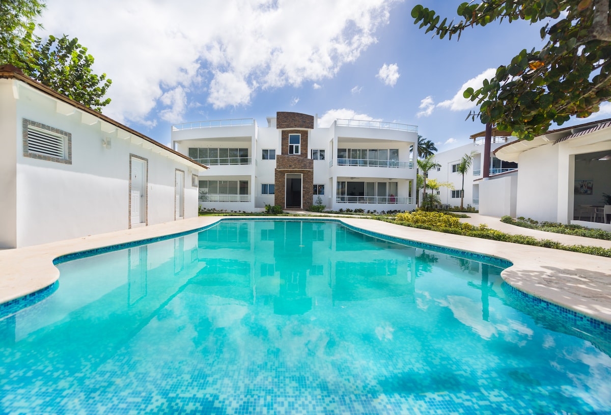 A serene swimming pool reflects the clear blue sky, surrounded by well-maintained gardens and the white exterior of the two-story building. Large windows provide a glimpse of the interior while palm trees gently sway in the background.