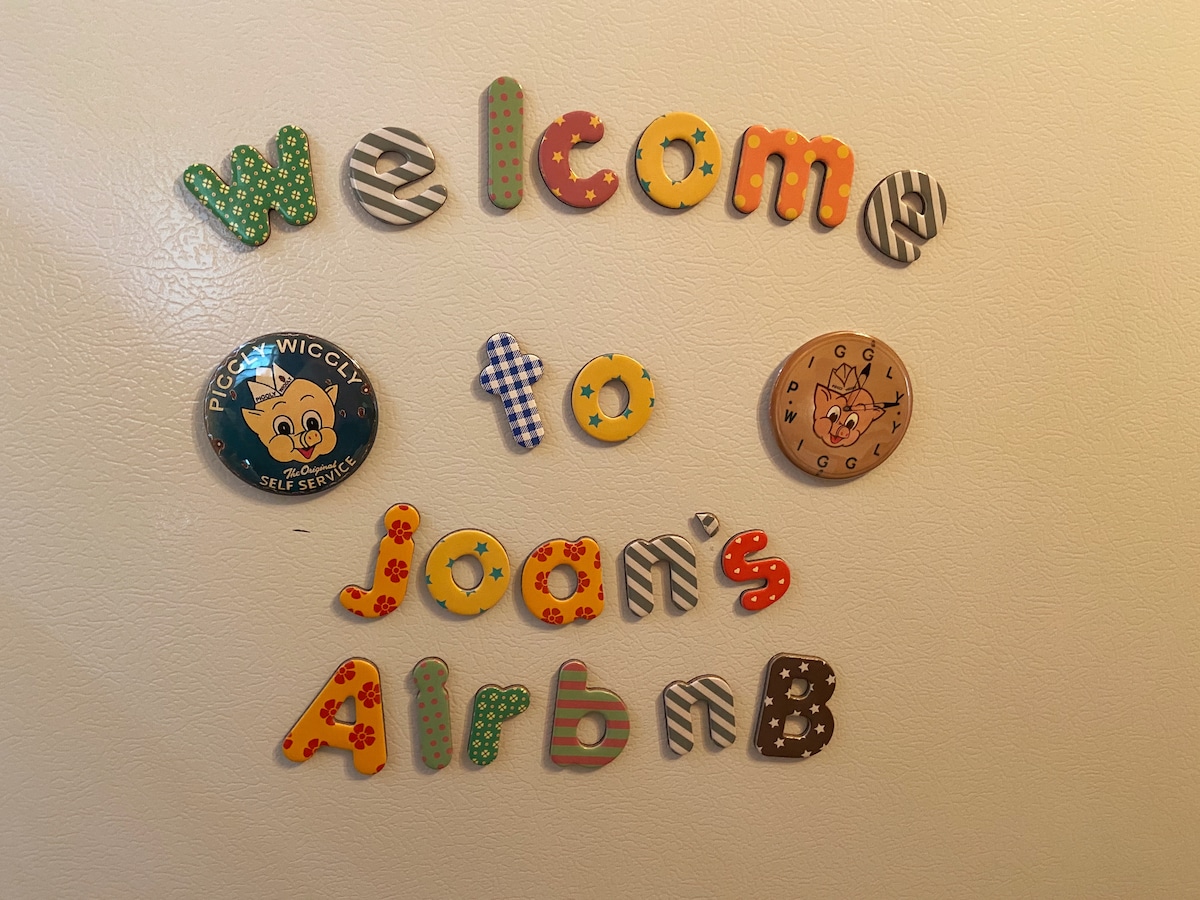 Colorful letters spell out 'Welcome to Joan's Airbnb' on a refrigerator door. Various designs and patterns adorn the letters, adding a playful touch. Two circular magnets featuring cartoon characters are positioned above and below the text.