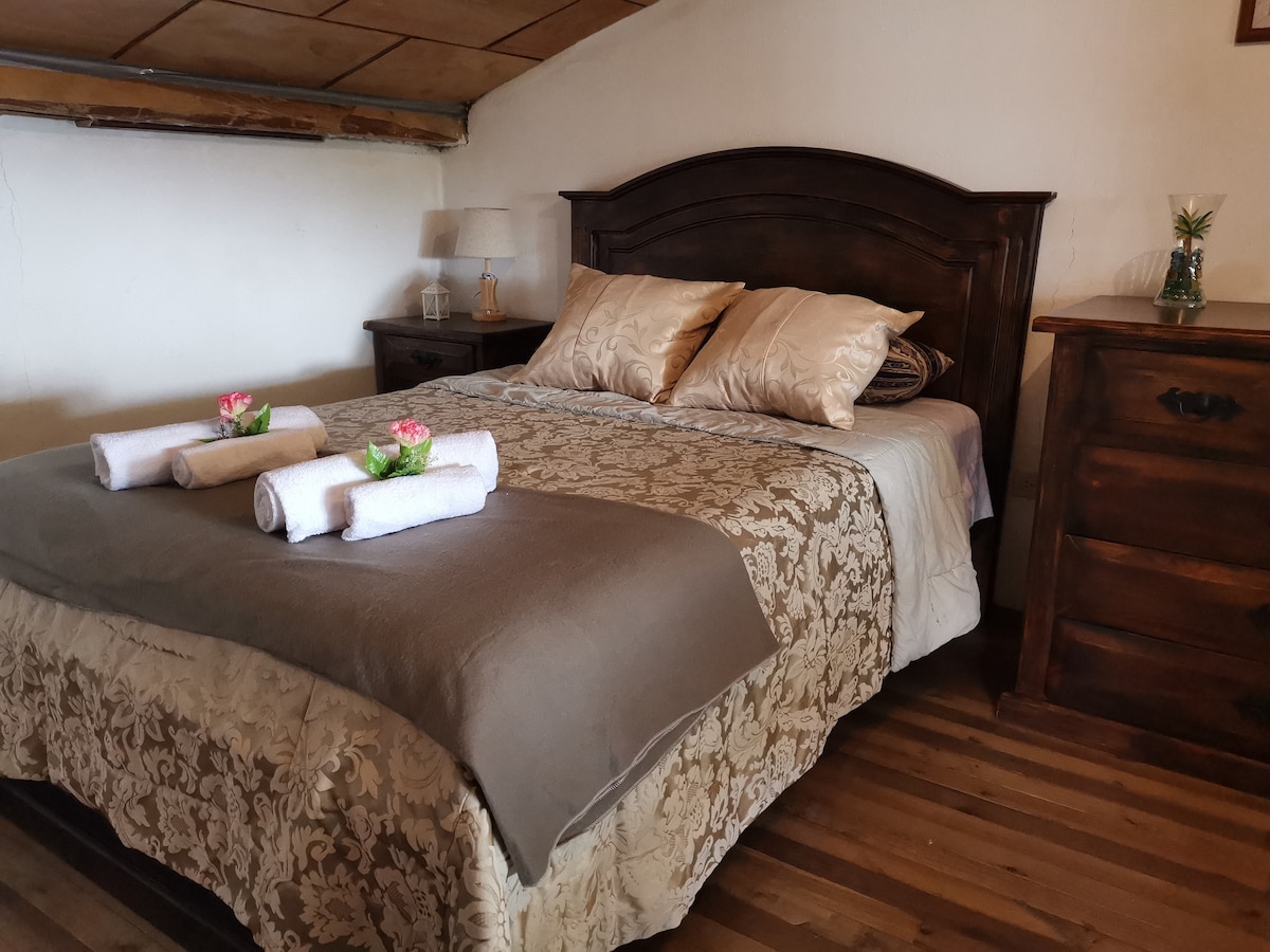 A spacious bed with elegant linens is centered in the room. Two decorative pillows and rolled towels are arranged on the bed's surface. A bedside table holds a lamp, while a wooden dresser complements the warm tones of the wooden flooring.