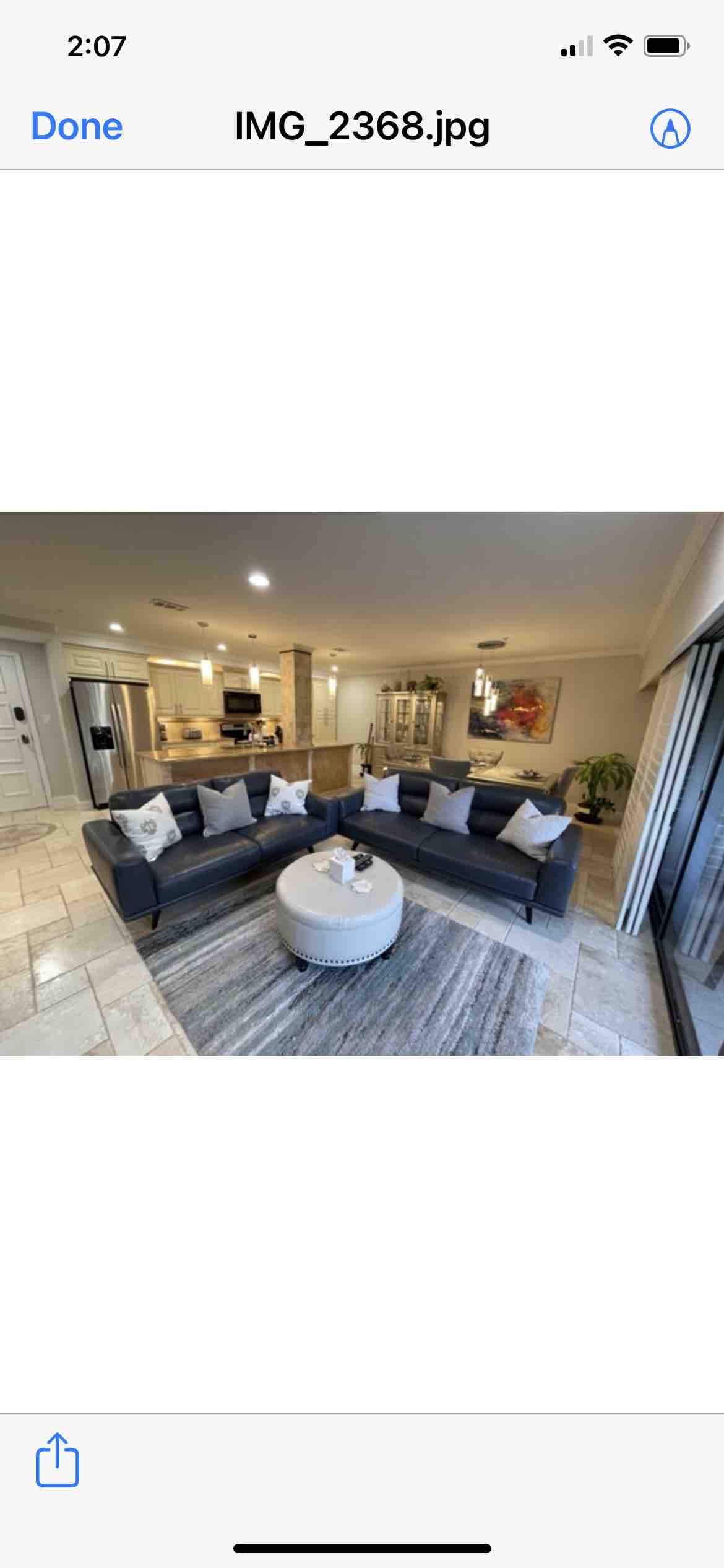 A modern living area features a spacious sectional sofa adorned with decorative pillows. A round coffee table is centered on a textured area rug. In the background, a fully equipped kitchen is visible, complemented by natural light flowing through large sliding glass doors.