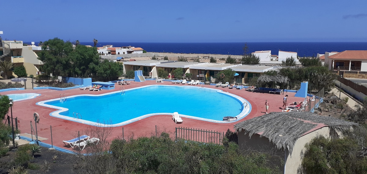 A large swimming pool is framed by sun loungers and umbrellas, set against a backdrop of blue ocean and clear skies. Lush greenery surrounds the pool area, enhancing the serene environment. The pool's shape offers various shallow and deep sections for relaxation.