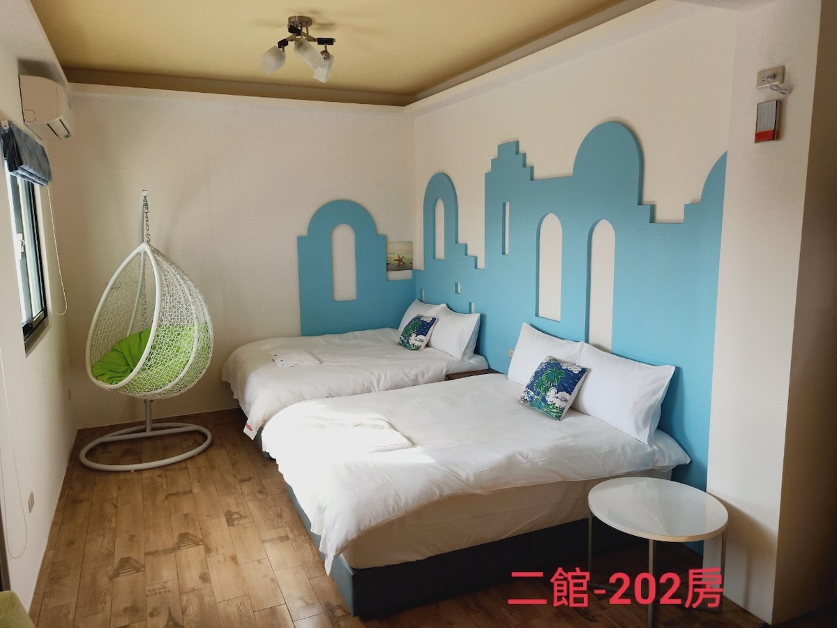 The room features two spacious beds with white linens, illuminated by natural light from a window. A unique blue mural with architectural motifs enhances the wall. A hanging chair adds a playful touch, while a small round table provides additional convenience.