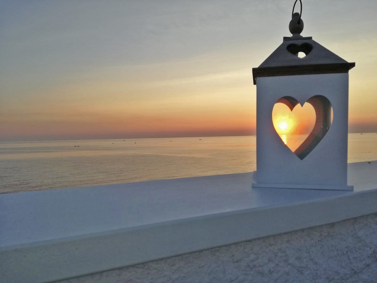 A decorative lantern with a heart-shaped cutout is positioned against a backdrop of a sunset over the calm sea. The warm colors of the sky reflect on the water's surface, with soft hues of orange, pink, and purple marking the horizon.