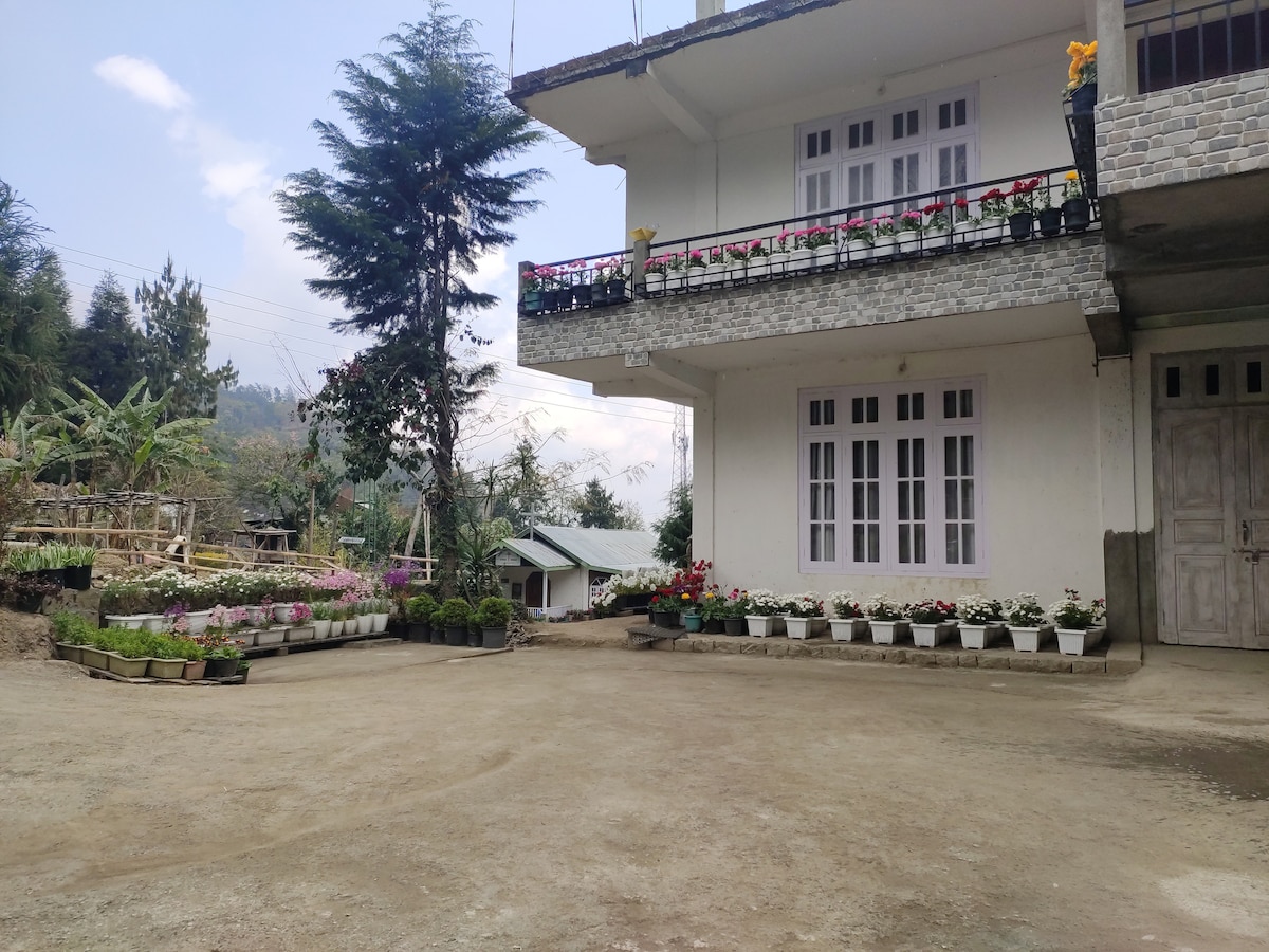 A cozy two-story building is positioned within a landscaped compound. Flower pots with various plants are arranged along the perimeter, while a pathway leads towards an outdoor space. The scene is enhanced by distant greenery, contributing to the tranquil atmosphere.