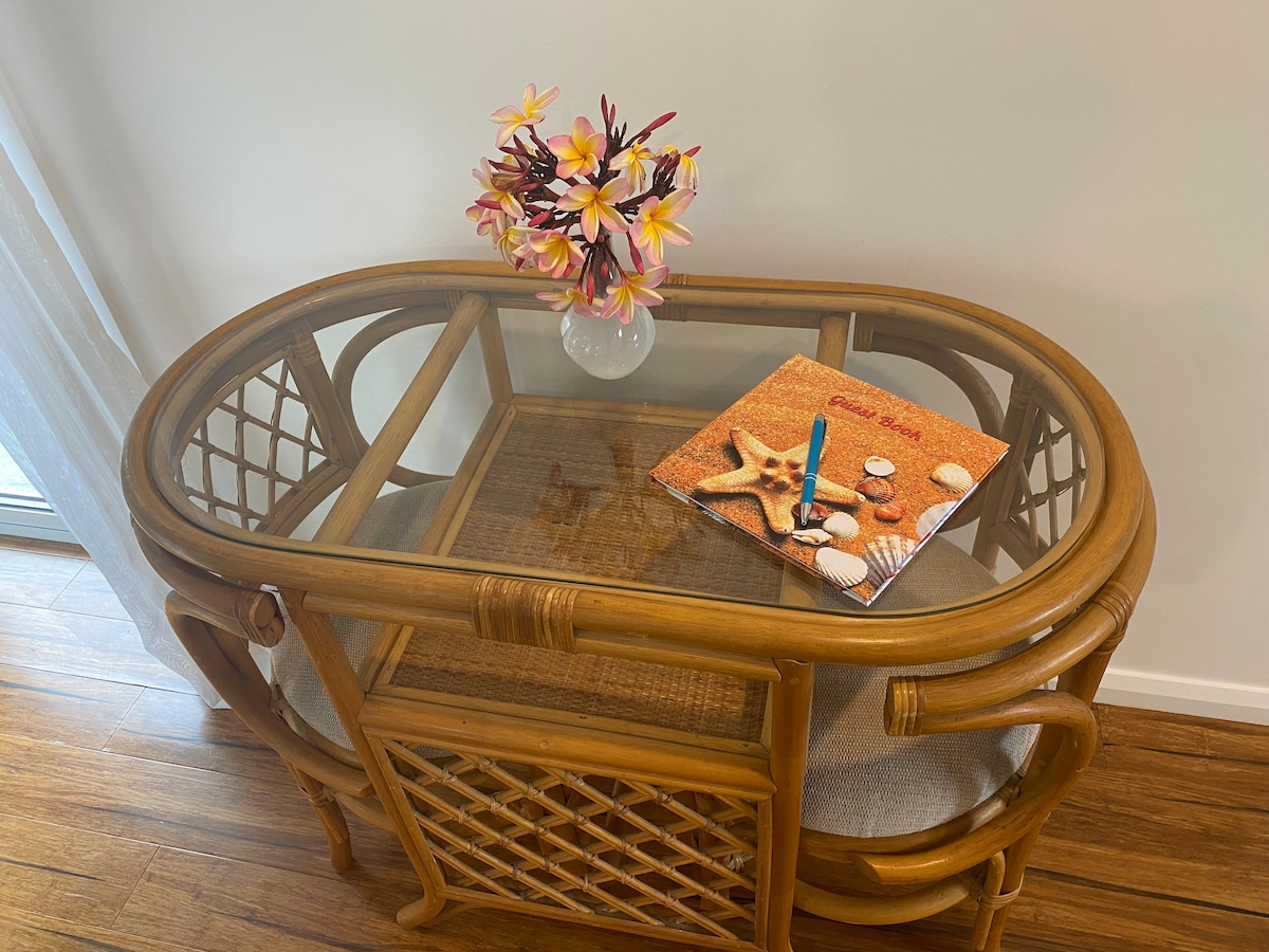 A round, wooden table with a glass top and woven design is positioned near a window. A vase containing colorful artificial flowers sits atop the table, alongside a magazine and a pen, creating a welcoming space for relaxation.