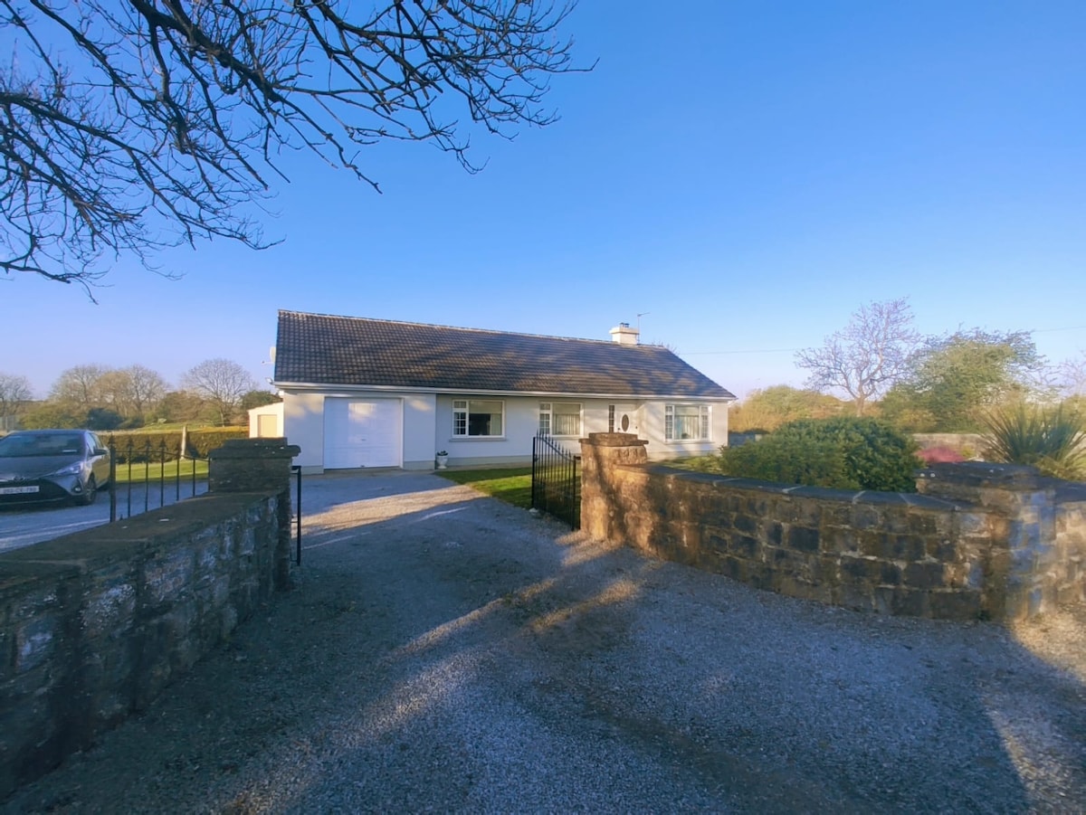 A single-story residence is depicted, featuring a sloped roof and large windows. The property is set against a clear blue sky, surrounded by greenery. A gravel driveway leads to the home, with a stone boundary wall and gated entrance creating a welcoming sense of arrival.