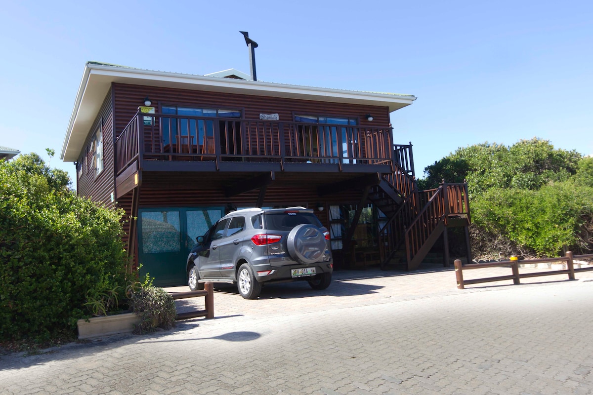 A rustic wooden cabin is visible with a deck overlooking the surroundings. A grey vehicle is parked in front, and a staircase leads to the upper entrance. Lush greenery surrounds the structure, providing a natural backdrop.