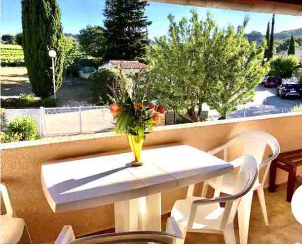A balcony area features a table with a vase of flowers at its center. Surrounding the table are white chairs, enhancing the outdoor dining space. Lush greenery and distant hills are visible beyond, providing a serene backdrop.