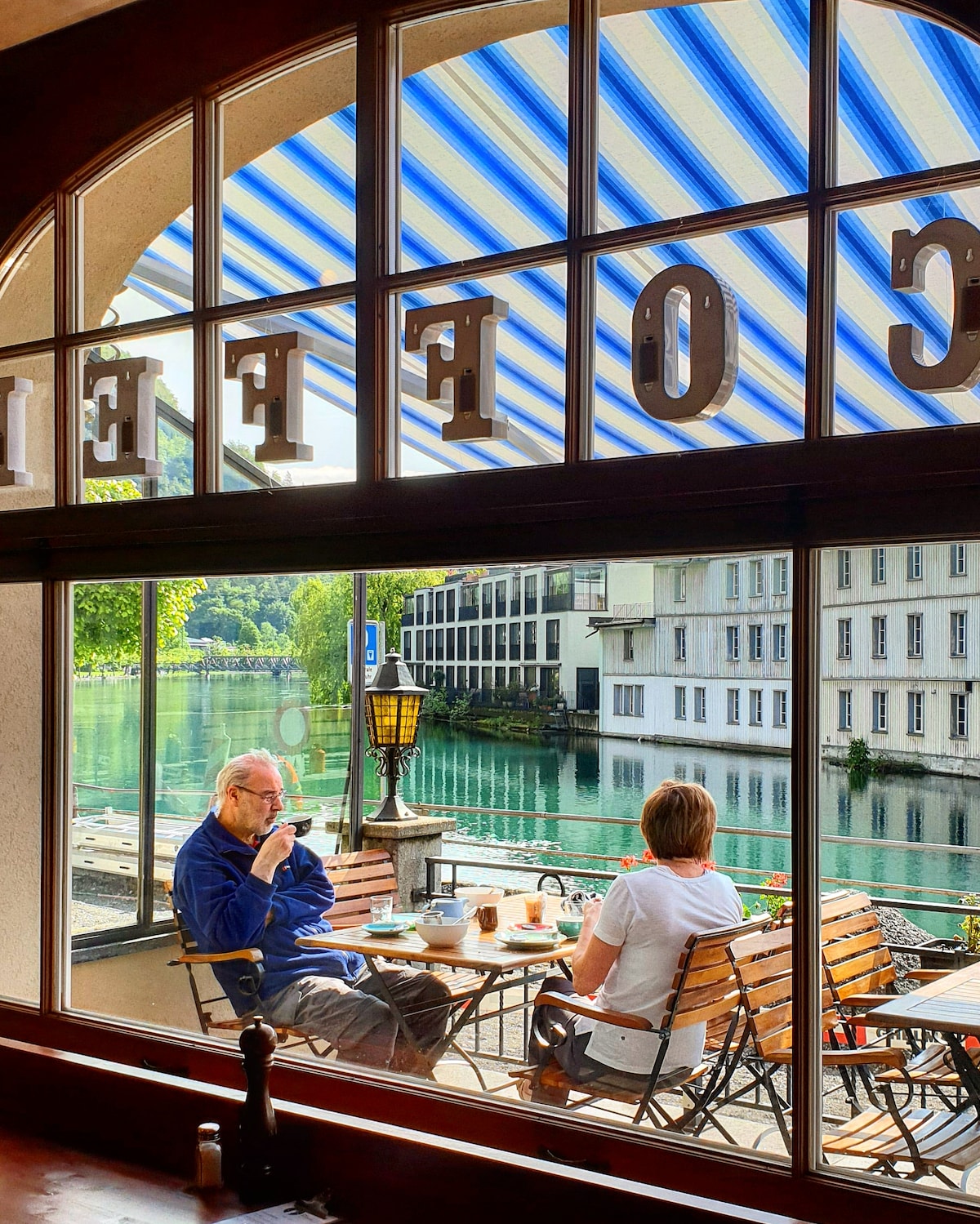 A café terrace is viewed through an arched window, where guests are seated at wooden tables with chairs. The serene River Aare reflects greenery, while the striped blue awning provides shade. Nearby buildings enhance the picturesque riverside atmosphere.