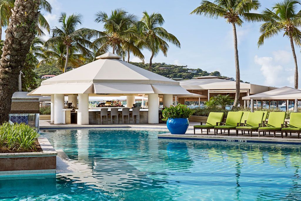 A quarter-acre pool is surrounded by lush greenery and palm trees, featuring a shaded cabana that offers seating and a bar area. Lounge chairs with bright cushions line the poolside, overlooking the tranquil waters and inviting atmosphere of the resort.