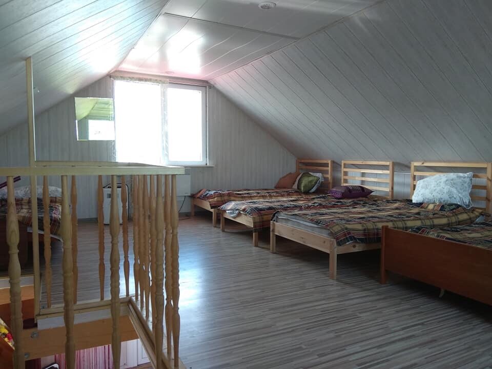 An attic space is revealed, featuring several wooden beds arranged with patterned bedding. Natural light enters through a large window, highlighting the wooden flooring and sloped ceiling. A cozy atmosphere is created with soft furnishings and a warm colour palette.