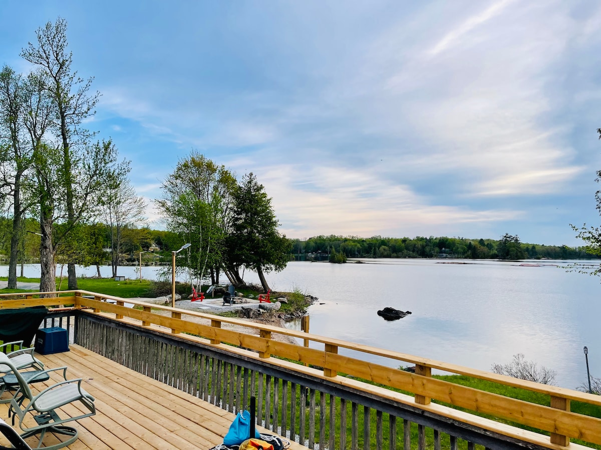 A spacious deck overlooks the serene waters of Stoney Lake, framed by trees and gentle slopes. Red kayaks are positioned on the shore, inviting exploration. Soft clouds drift across the sky, reflecting the tranquil ambiance of the surrounding landscape.