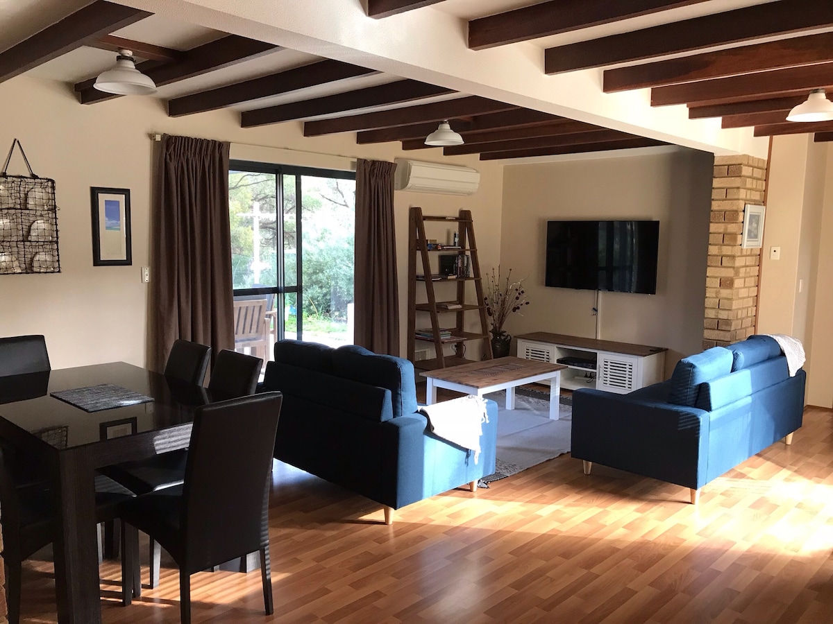 An open-plan living and dining area is featured, showcasing two blue sofas and a dining table with black chairs. Large windows provide natural light, and a television is mounted on a brick wall. Wooden beams create an inviting ceiling structure.