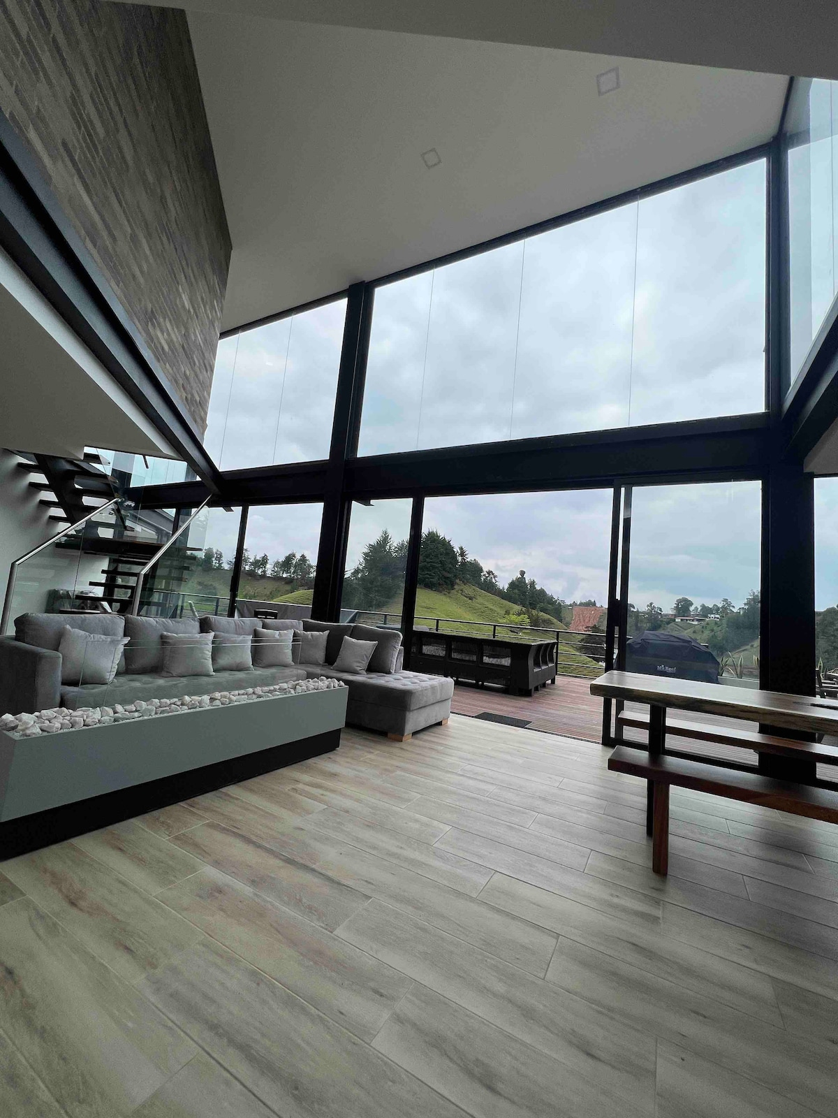 A spacious living area is characterized by large glass walls allowing natural light and scenic views. A comfortable sectional sofa is arranged around a sleek fireplace, and a dining table is positioned nearby. Polished wooden floors enhance the contemporary design.