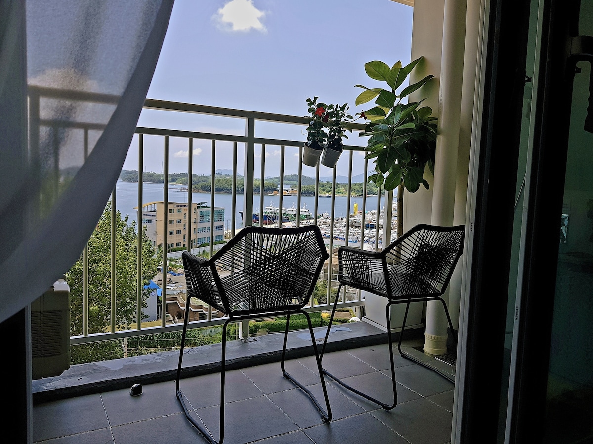 A serene balcony view showcases two black wicker chairs, positioned beside a small potted plant. Beyond the railing, a scenic waterfront is visible, with blue water and buildings in the distance. The soft clouds float in a clear sky, enhancing the tranquil setting.