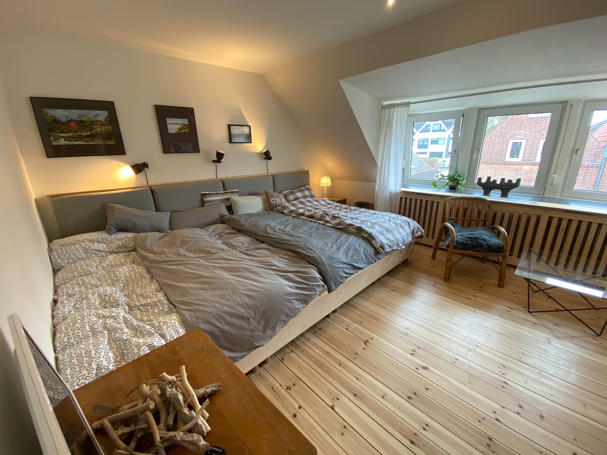 A spacious bedroom features two large beds arranged neatly, each adorned with muted linens. Natural light fills the room through large windows, illuminating simple decorations on the walls and providing a calming atmosphere. A wooden table and a few minimalist chairs complete the setup.