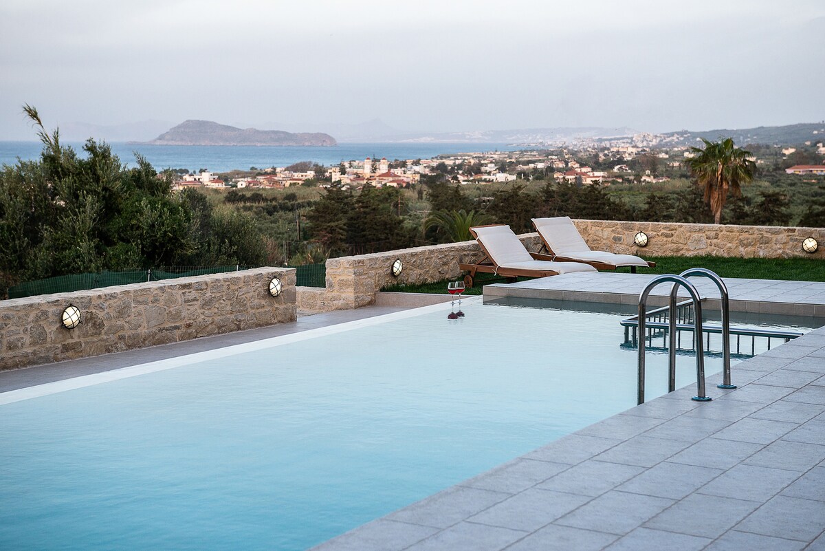 A serene outdoor pool area is featured, with clear water reflecting the sky. Two sun loungers are positioned at the edge, overlooking a picturesque view of the sea and distant land. Olive trees and Mediterranean vegetation surround the area, enhancing the tranquil atmosphere.