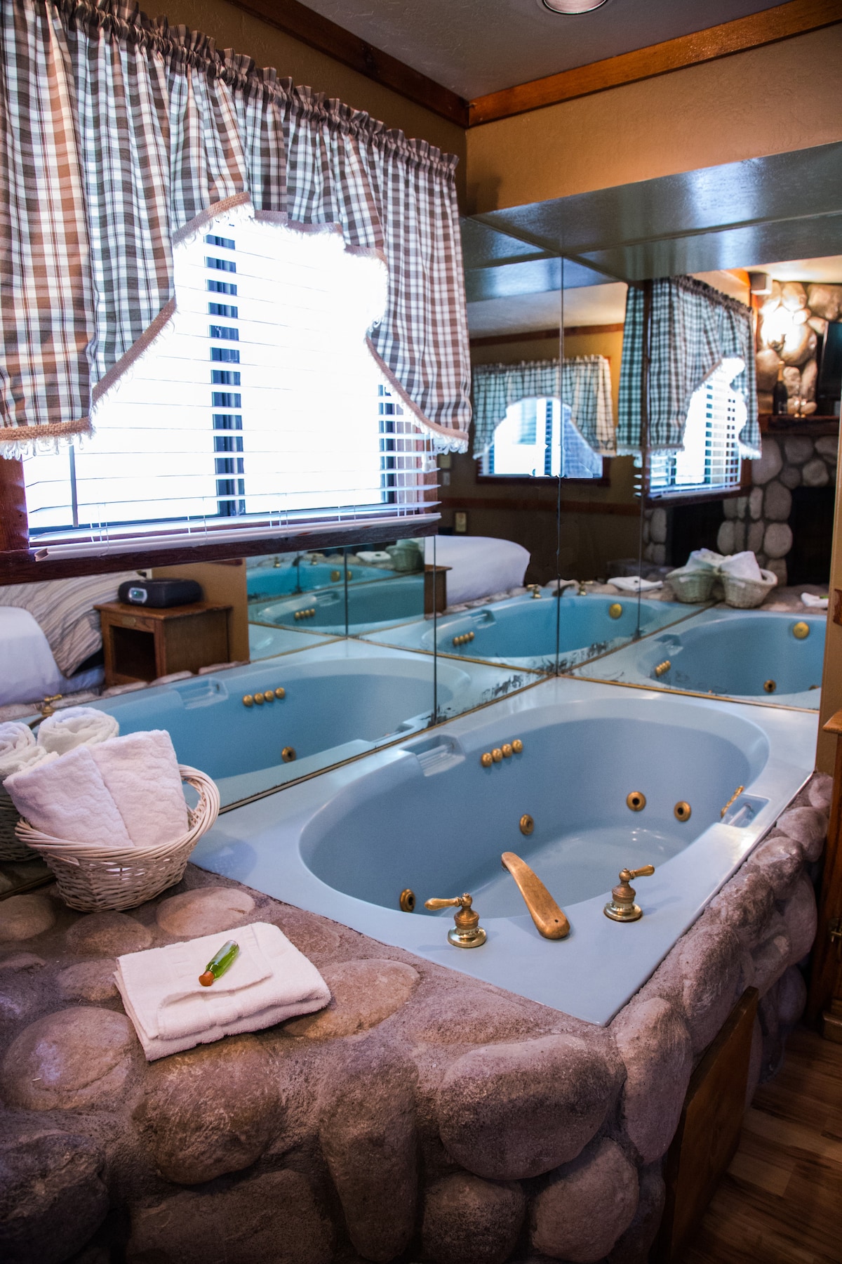 A river-rock jetted bath is positioned beside a mirror-covered wall, reflecting the warm lighting of the space. A basket of neatly rolled towels is arranged on the counter, with a stone surface surrounding the bath. Large windows with checked valances offer natural light.