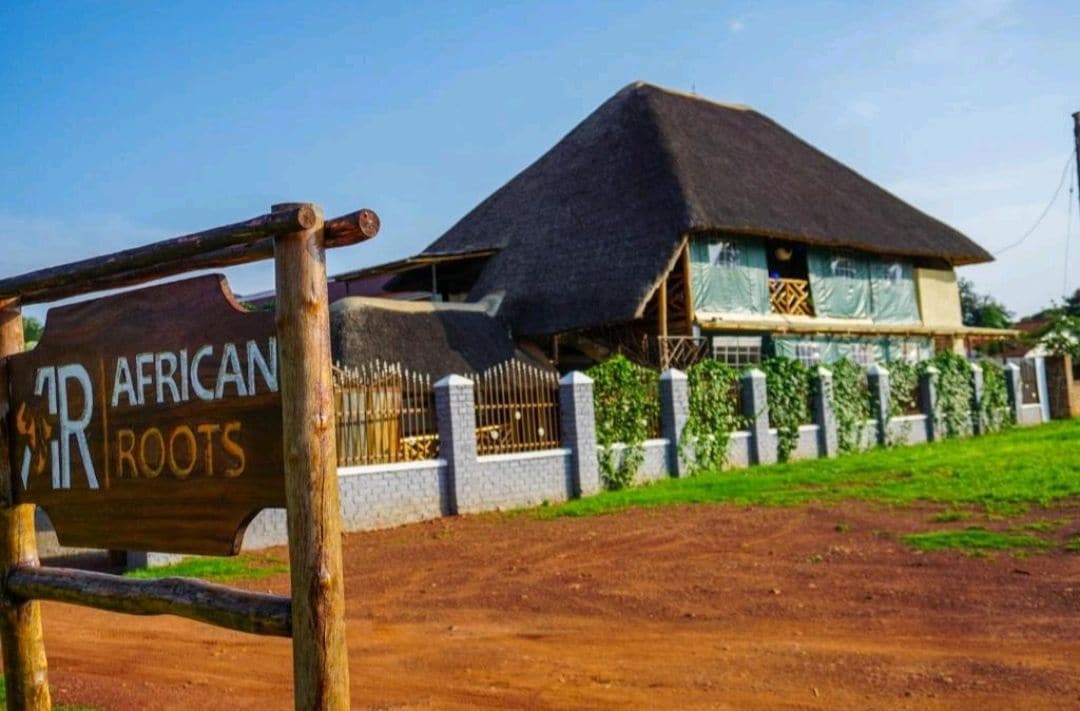The exterior of the African Roots Hotel is showcased, featuring a thatched roof and wooden signage. Lush greenery surrounds the property, with a clear blue sky overhead. The entrance is framed by a rustic wooden fence, providing an inviting view of the hotel.