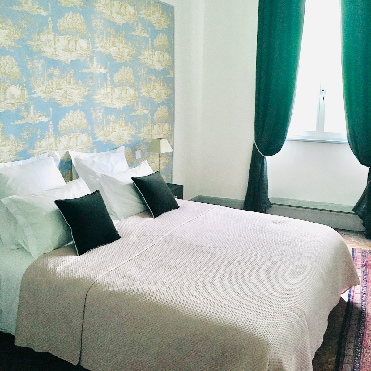 A cozy bedroom features a spacious bed adorned with neutral bedding and accent pillows. The light blue wallpaper with a nature motif complements the room. Sheer curtains frame the window, allowing natural light to softly illuminate the space.