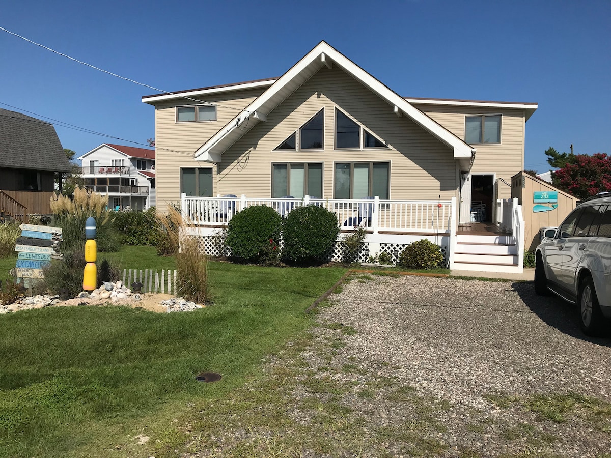 The exterior of a spacious two-story vacation home is shown, featuring a wide porch with white railings and large windows. A well-maintained lawn with decorative plants lines the front, and gravel leads to the driveway. Clear blue skies complement the inviting atmosphere.