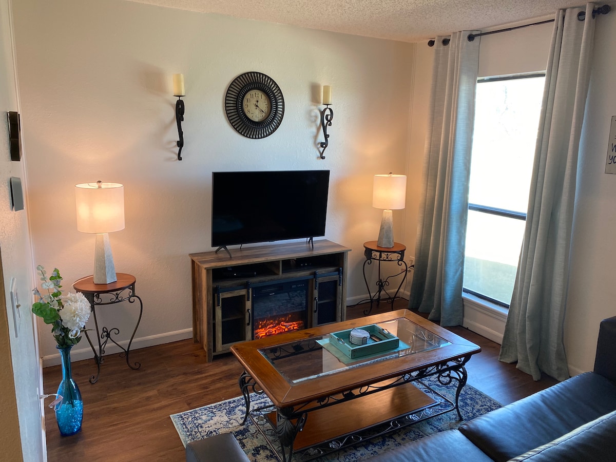 A cozy living room displays a media console beneath a clock, with an electric fireplace integrated within. Two lamps on side tables provide soft lighting. A coffee table sits at the center, accompanied by a decorative tray. Large windows offer natural light and a view of the outdoors.