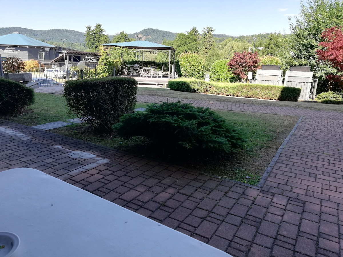 A view of the landscaped garden features neatly trimmed hedges and grass, with a stone path leading towards a gazebo area equipped with tables and chairs. Surrounding trees provide a serene backdrop, while distant hills create a calming atmosphere.