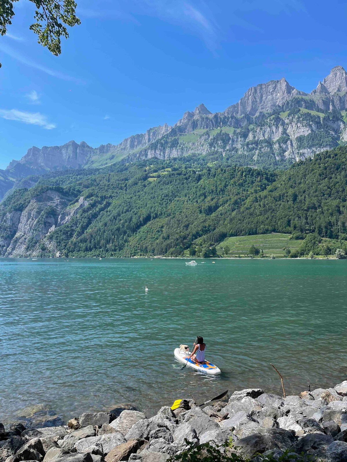 A person is seen on a paddleboard in clear waters, surrounded by mountains that rise steeply in the background. The shoreline is characterized by smooth stones leading to the water, with green hills cascading towards the lake.