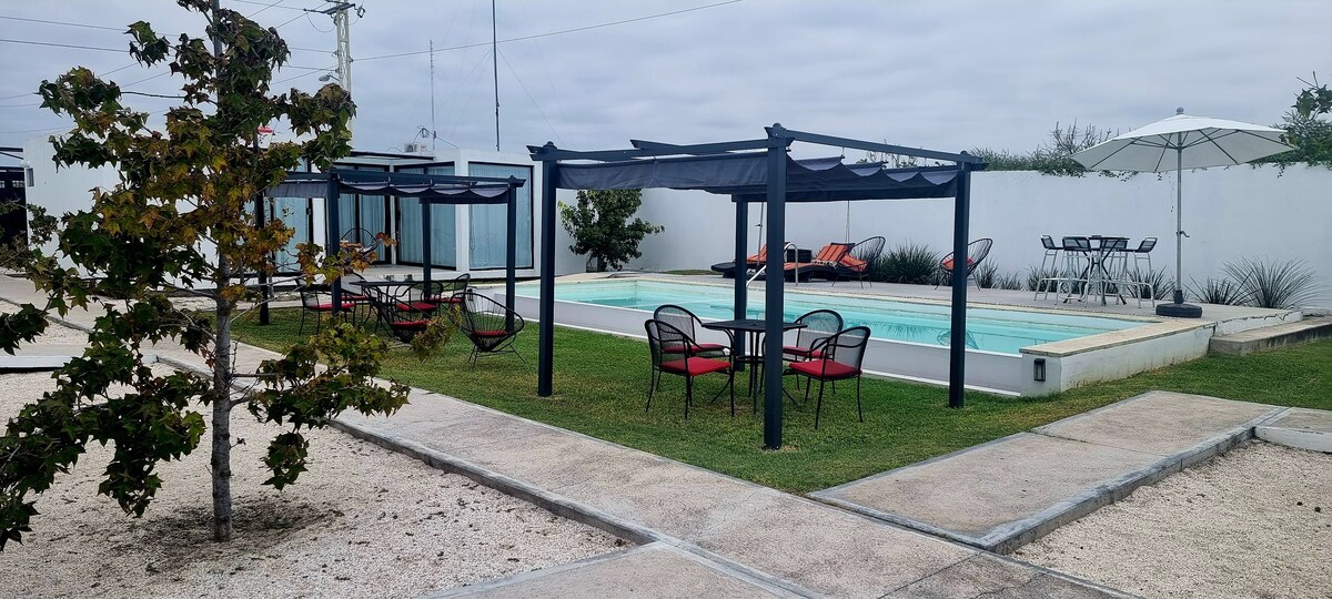 An outdoor area features a solar-heated pool surrounded by lounge chairs and shaded seating areas under pergolas. Table and chair sets are nearby, and a patio umbrella offers additional shade. Green grass and decorative trees enhance the inviting, serene environment.