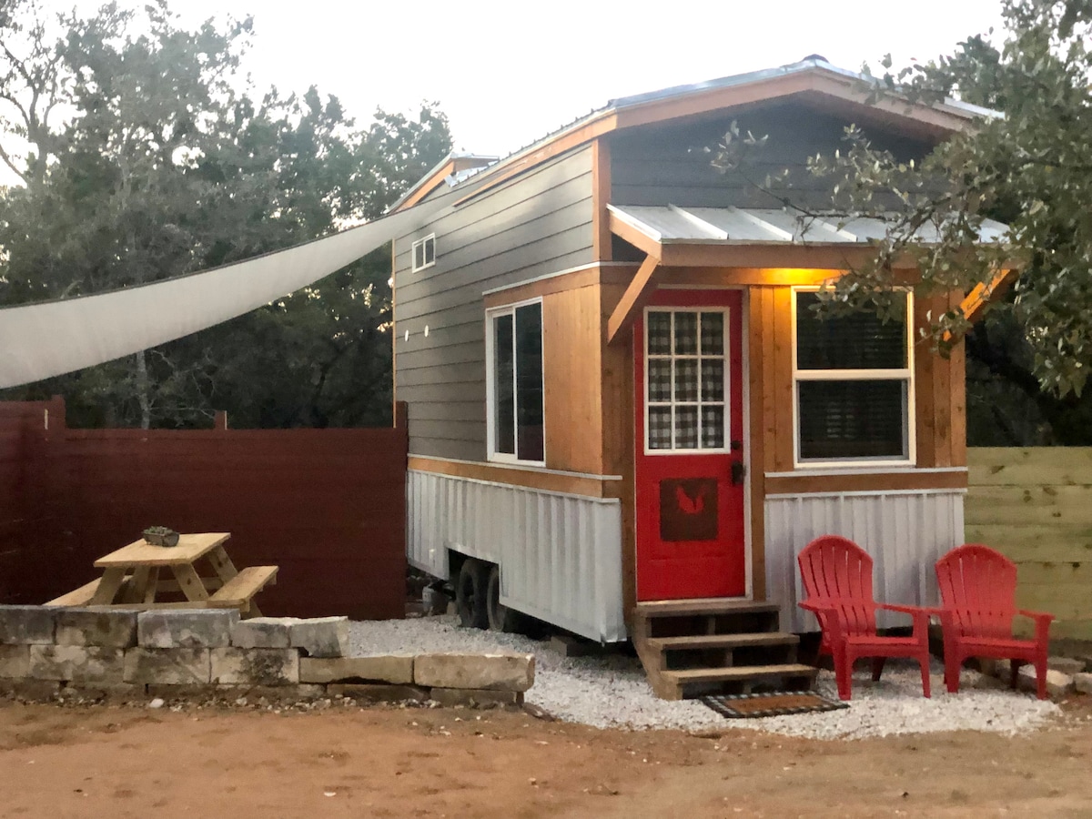 The tiny home features a charming exterior with wooden accents and a bright red door. Two red chairs are positioned invitingly on a gravel area, alongside a picnic table. A shaded canopy provides protection from the sun, while surrounding trees add natural beauty.
