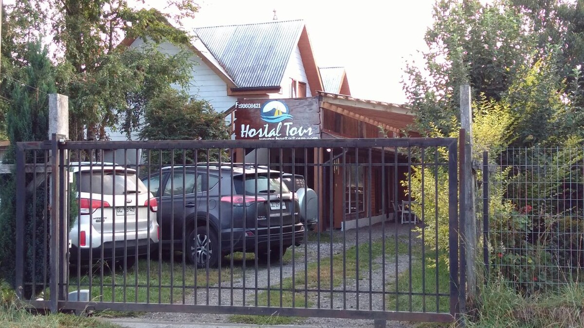 The image showcases the entrance to Hostal Tour Pucon, featuring a gated entrance with a wooden sign displaying the hostel's name. Several parked vehicles are visible in the driveway, alongside a gravel path leading towards the building, which is surrounded by greenery.